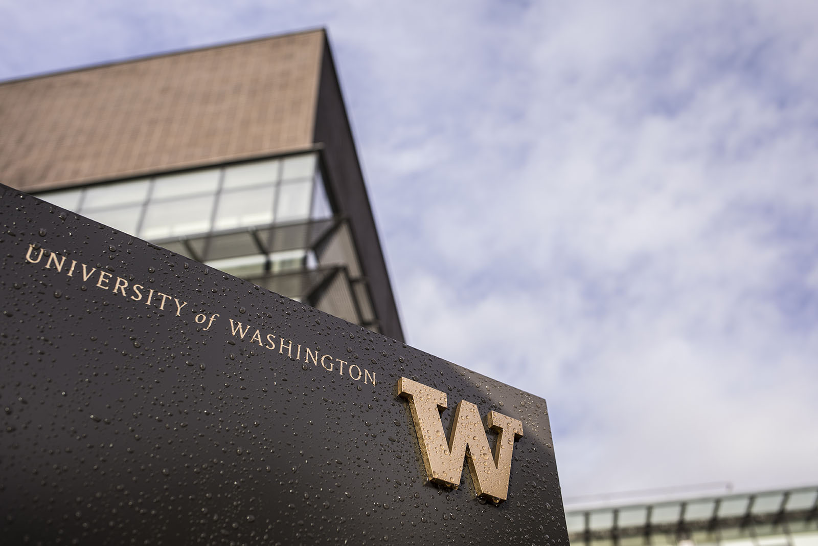 A rain-covered University of Washington monument sign with a gold W, with a campus building and cloudy sky behind it.